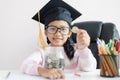 Little Asian girl wearing graduate hat putting the coin into clear glass jar piggy bank and smile with happiness for money saving Royalty Free Stock Photo