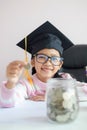 Little Asian girl wearing graduate hat putting the coin into clear glass jar piggy bank and smile with happiness for money saving Royalty Free Stock Photo