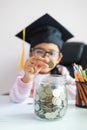 Little Asian girl wearing graduate hat putting the coin into clear glass jar piggy bank and smile with happiness for money saving Royalty Free Stock Photo