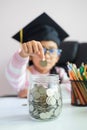 Little Asian girl wearing graduate hat putting the coin into clear glass jar piggy bank and smile with happiness for money saving Royalty Free Stock Photo