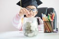 Little Asian girl wearing graduate hat putting the coin into clear glass jar piggy bank and smile with happiness for money saving Royalty Free Stock Photo