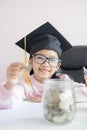 Little Asian girl wearing graduate hat putting the coin into clear glass jar piggy bank and smile with happiness for money saving Royalty Free Stock Photo