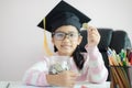 Little Asian girl wearing graduate hat putting the coin into clear glass jar piggy bank and smile with happiness for money saving Royalty Free Stock Photo