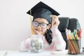 Little Asian girl wearing graduate hat putting the coin into clear glass jar piggy bank and smile with happiness for money saving Royalty Free Stock Photo