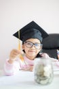 Little Asian girl wearing graduate hat putting the coin into clear glass jar piggy bank and smile with happiness for money saving Royalty Free Stock Photo