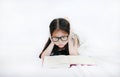 Little Asian kid girl wearing glasses reading hardcover book lying on bed against white background Royalty Free Stock Photo