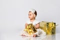 Little babe in dress and headband, barefoot. She posing with two golden gift boxes, sitting on floor  on white. Close up Royalty Free Stock Photo