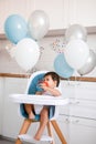 Little baby boy sitting in blue high chair at home on white kitchen and drinking water from sippy cup on background with balloons Royalty Free Stock Photo
