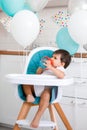 Little baby boy sitting in blue high chair at home on white kitchen and drinking water from sippy cup on background with balloons Royalty Free Stock Photo