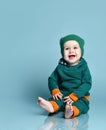 Little baby boy in stylish casual jumpsuit, cap and barefoot sitting on floor and smiling over blue wall background Royalty Free Stock Photo