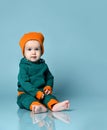 Little baby boy in stylish casual jumpsuit, cap and barefoot sitting on floor and smiling over blue wall background Royalty Free Stock Photo