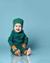 Little baby boy in stylish casual jumpsuit, cap and barefoot sitting on floor and smiling over blue wall background Royalty Free Stock Photo