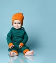 Little baby boy in stylish casual jumpsuit, cap and barefoot sitting on floor and smiling over blue wall background Royalty Free Stock Photo