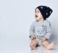 Little baby boy toddler in grey casual jumpsuit, black cap with stars and barefoot sitting on floor, smiling and looking aside Royalty Free Stock Photo