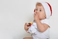 Little baby in christmas hat looking away with astonishing look, covering his mouth with tiny fingers, holding plastic dog toy, Royalty Free Stock Photo