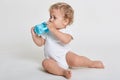 Little baby drinking water from blue bottle, looking aside while sitting on floor, posing barefoot and dresses bodysuit, cute Royalty Free Stock Photo