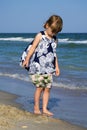 Little baby in a flowery dress is standing on the sea shore with her head down and looking like a sea wave washes her feet