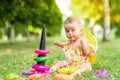 Little baby girl 7 months old sitting on the green grass in a yellow dress and hat and playing with a pyramid, early development Royalty Free Stock Photo