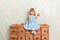 Little baby girl sitting cross-legged on chest of drawers holding tangerine in hand and pointing down with index finger Royalty Free Stock Photo