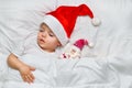 Little baby sleeping on white linen in the Santa hat with his toy Santa Claus.