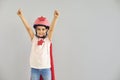 Little baby superhero in a helmet smiles raised his hands while standing against a gray background.