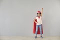 Little baby superhero in a helmet smiles raised his hands while standing against a gray background.