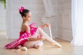 Little ballerina in a bright pink tutu sits on the floor in a bright hall and puts on pointe shoes