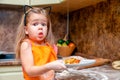 Little beautiful cute girl in orange apron smiling and making homemade pizza, roll the dough at home kitchen. Concept happy family Royalty Free Stock Photo