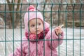 Little beautiful girl baby behind the fence, grid locked in a cap and a jacket with sad emotion on his face