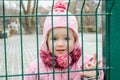 Little beautiful girl baby behind the fence, grid locked in a cap and a jacket with sad emotion on his face