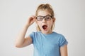 Little beautiful girl with bright blue eyes and blonde hair posing with opened mouth, holding her glasses with hand Royalty Free Stock Photo