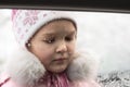 Little beautiful girl dressed in a warm pink jacket and hat, in winter, looks at snowflakes on a car window Royalty Free Stock Photo