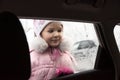 Little beautiful girl dressed in a warm pink jacket and hat, in winter, looks at snowflakes on a car window Royalty Free Stock Photo