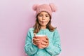 Little beautiful girl holding pink mug wearing cute wool hat making fish face with mouth and squinting eyes, crazy and comical