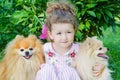 Little Beautiful Girl Hugging German And Pomeranian On Green Grass. Child With Pet In Nature. Friendship Between Dog And Human. Ha