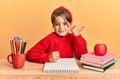 Little beautiful girl sitting on classroom desk smiling with happy face looking and pointing to the side with thumb up Royalty Free Stock Photo