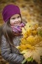 Little beautiful girl stands with a bouquet of maple orange leaves in the autumn park. She looks away and smiles. Dressed in a Royalty Free Stock Photo