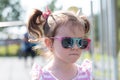 Little beautiful sad girl in sun-protective glasses with pigtails stands on the street Royalty Free Stock Photo