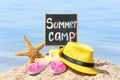 Little blackboard with text SUMMER CAMP, hat and flip flops on beach near sea Royalty Free Stock Photo