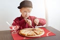 A little blond boy with blue eyes wearing red shirt and stylish cap sitting in cosy cafe eating delicious pizza and ice-cream look Royalty Free Stock Photo