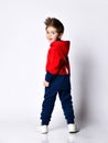 Little blond boy in blue and red tracksuit, sneakers, sunglasses. Smiling turning back, showing thumb up, posing isolated on white Royalty Free Stock Photo