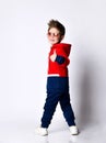 Little blond boy in blue and red tracksuit, sneakers, sunglasses. Smiling turning back, showing thumb up, posing isolated on white Royalty Free Stock Photo