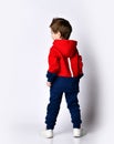 Little blond boy in blue and red tracksuit, sneakers, sunglasses. Smiling turning back, showing thumb up, posing isolated on white Royalty Free Stock Photo