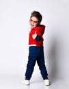 Little blond boy in blue and red tracksuit, sneakers, sunglasses. Smiling turning back, showing thumb up, posing isolated on white Royalty Free Stock Photo