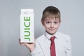 Little blond boy holds and shows a big white carton juice package. White shirt and red tie. Light background