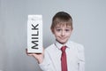 Little blond boy holds and shows a big white carton milk package. White shirt and red tie. Light background Royalty Free Stock Photo