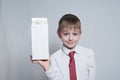 Little blond boy holds and shows a big white carton package. White shirt and red tie. Light background Royalty Free Stock Photo