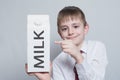 Little blond boy holds and shows the index finger on a large white carton milk package. White shirt and red tie. Light background