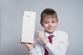 Little blond boy holds and shows the index finger on a large white carton package. White shirt and red tie. Light background Royalty Free Stock Photo