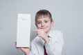 Little blond boy holds and shows the index finger on a large white carton package. White shirt and red tie. Light background Royalty Free Stock Photo